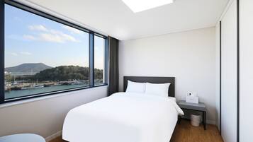 A modern hotel room with a large bed, a bedside table, a trash bin, and a view of a harbor with boats and hills.
