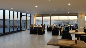 Un restaurante moderno con amplios ventanales que ofrecen vistas a la ciudad y al agua. Las mesas están ordenadas con sillas.