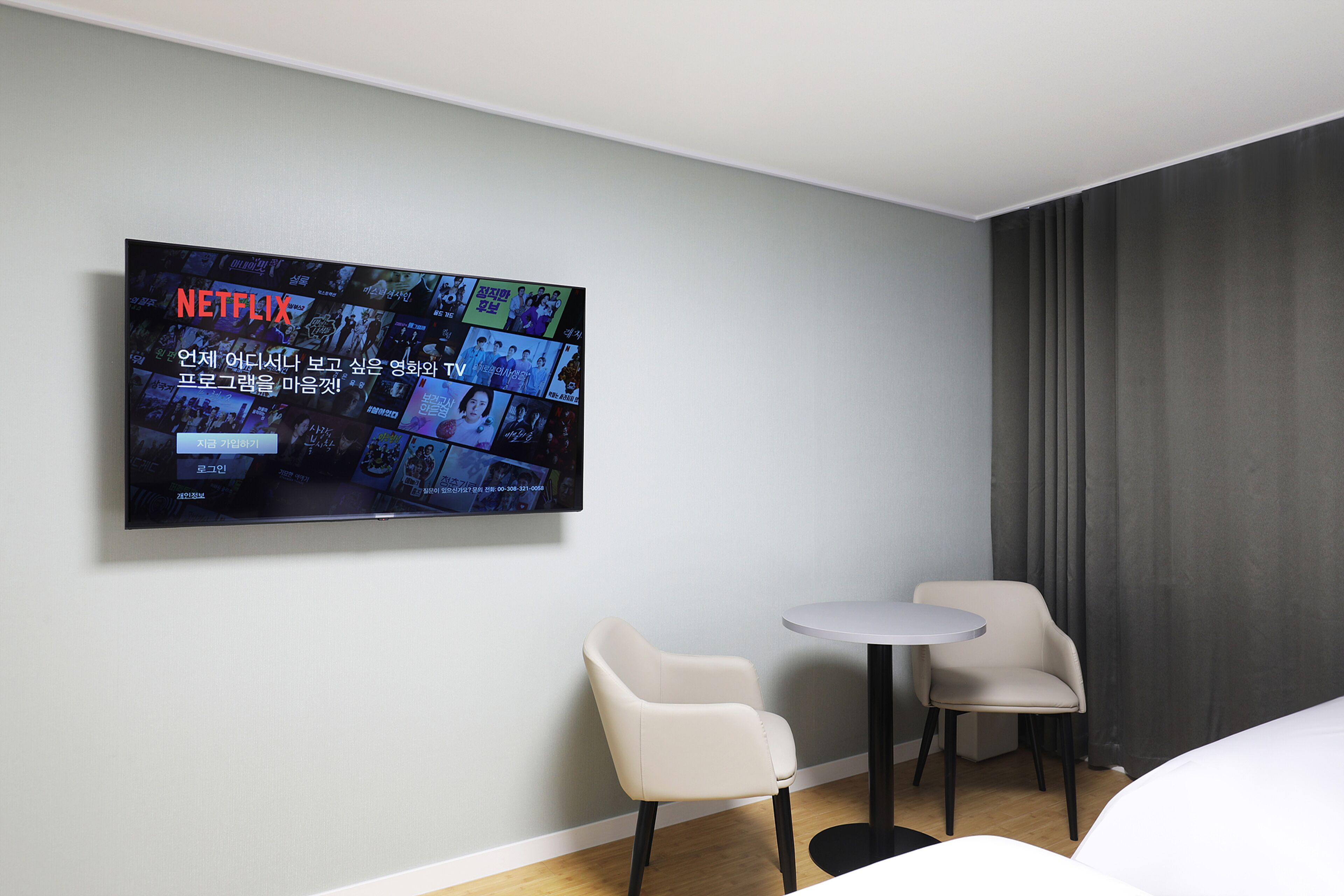 A modern hotel room with a large flat-screen TV mounted on the wall displaying a Netflix interface. There are two chairs and a small round table.