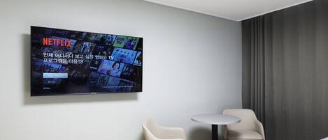 A modern hotel room with a large flat-screen TV mounted on the wall displaying a Netflix interface. There are two chairs and a small round table.