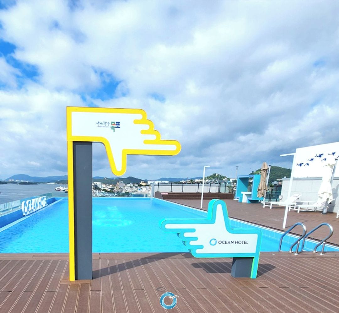 A pool area with a sign for the Ocean Hotel, a deck with lounge chairs, and a view of the sea and distant land.