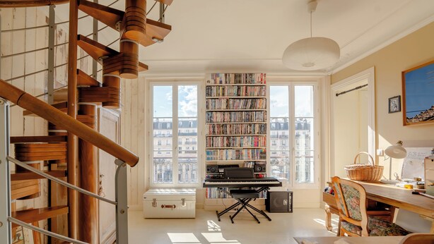 Une pièce avec un escalier en bois, une bibliothèque remplie de livres, un bureau avec un clavier et une fenêtre donnant sur des immeubles.