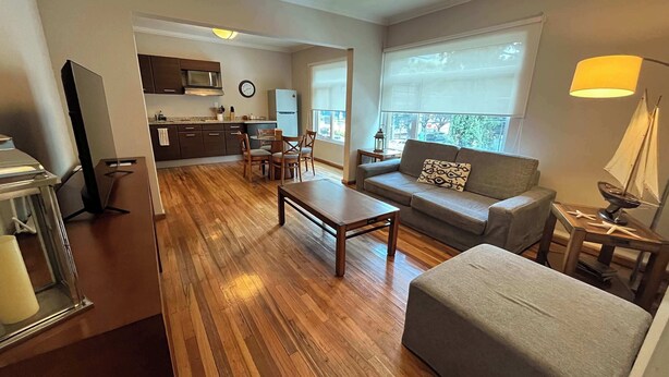A modern living room with a wooden floor, a sofa, a coffee table, and a TV. There is a kitchen area with a microwave and cabinets. - Arquimedes Deco - Colourful 1BR in Polanco (Mexico City)