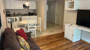 Uma sala moderna com um sofá marrom, uma mesa de jantar com cadeiras brancas, uma cozinha com armários brancos e uma TV sobre um suporte branco.