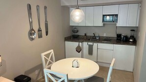 Cozinha compacta com armários brancos, micro-ondas e pia. Há uma mesa de jantar com cadeiras e um lustre.