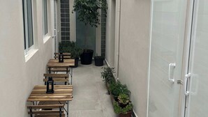 Un couloir étroit bordé d’une rangée de tables et de bancs en bois, de plantes en pot et d’une porte vitrée sur la droite.