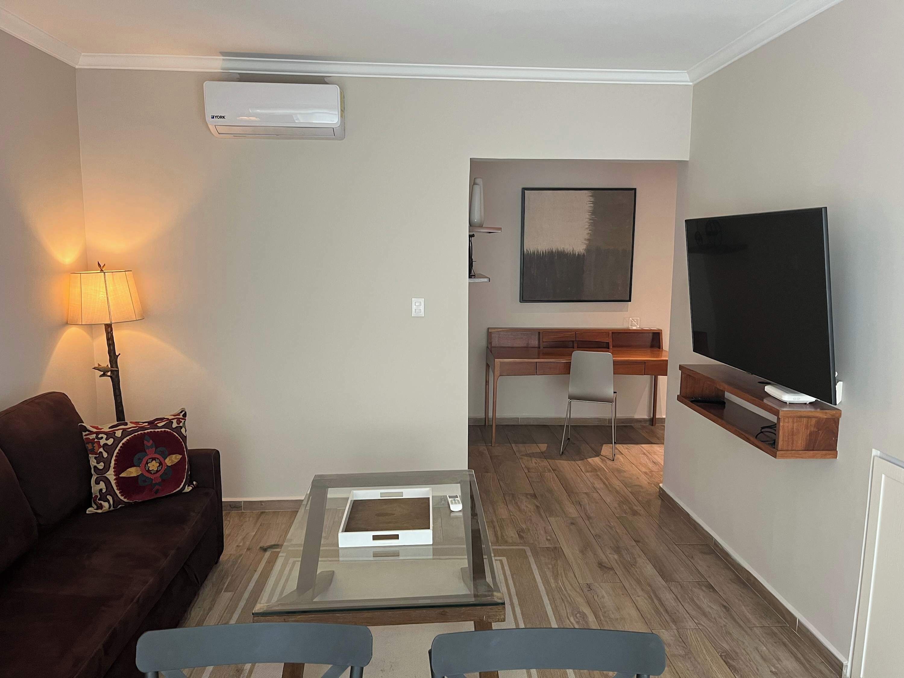 A modern living room with a glass coffee table, a sofa, a TV, and a desk with a chair.