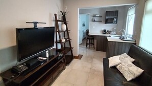 Ein modernes Wohnzimmer mit Flachbildfernseher, einer Holz-Unterhaltungseinheit und einem dunklen Sofa mit dekorativen Kissen. Im Hintergrund ist eine Küchenzeile mit Mikrowelle und Spüle erkennbar.