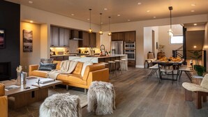 A modern living room with a sofa, coffee table, and ottoman, a kitchen with dark cabinets and stainless steel appliances, and a dining area with a table and chairs. - Summit + Sky (Bozeman)