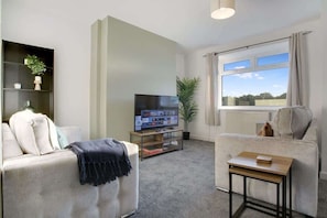 A living room with a sofa, a TV, a small table, a plant, and a window with a view.