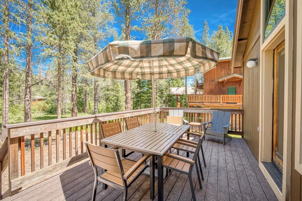 An outdoor deck with a wooden table and chairs, a checkered umbrella, and a view of a forest. - Rustic Retreat by Tahoe Getaways - 3 BR Dog Friendly in Tahoe Donner w/ HOA Beach, Gym, Pool (Truckee)