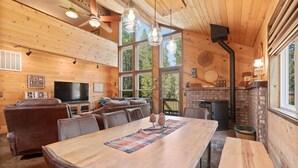 A cozy cabin interior with a large dining table, leather sofas, a fireplace, and a television.