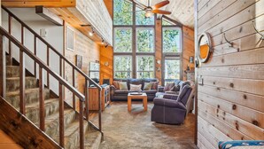 A cozy cabin interior with a wooden staircase, a living area with a sofa and armchairs, and large windows offering a view of the forest.