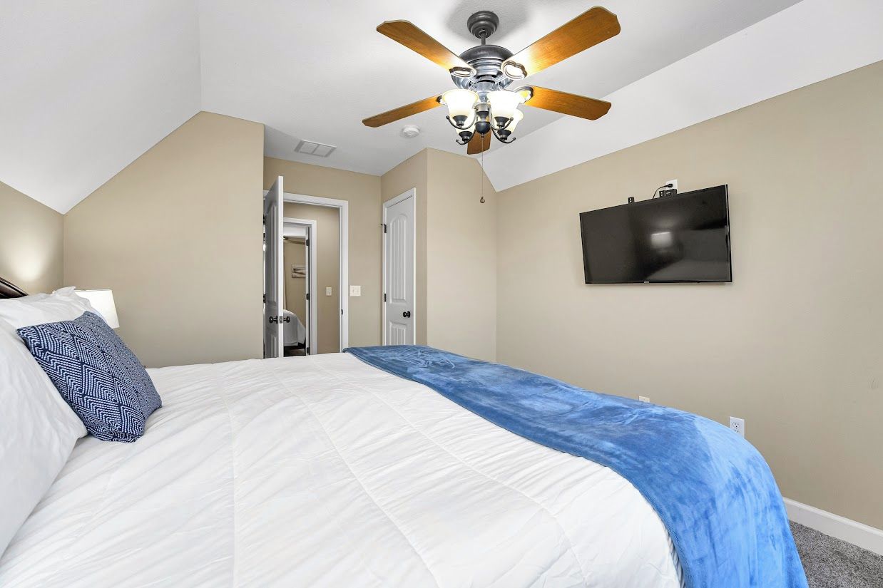 A bedroom with a bed, a ceiling fan, a television on the wall, and a door leading to another room.