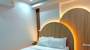 Cama bem arrumada com lençóis brancos e duas toalhas dobradas, cabeceira de madeira com iluminação suave e um criado-mudo simples com um telefone.