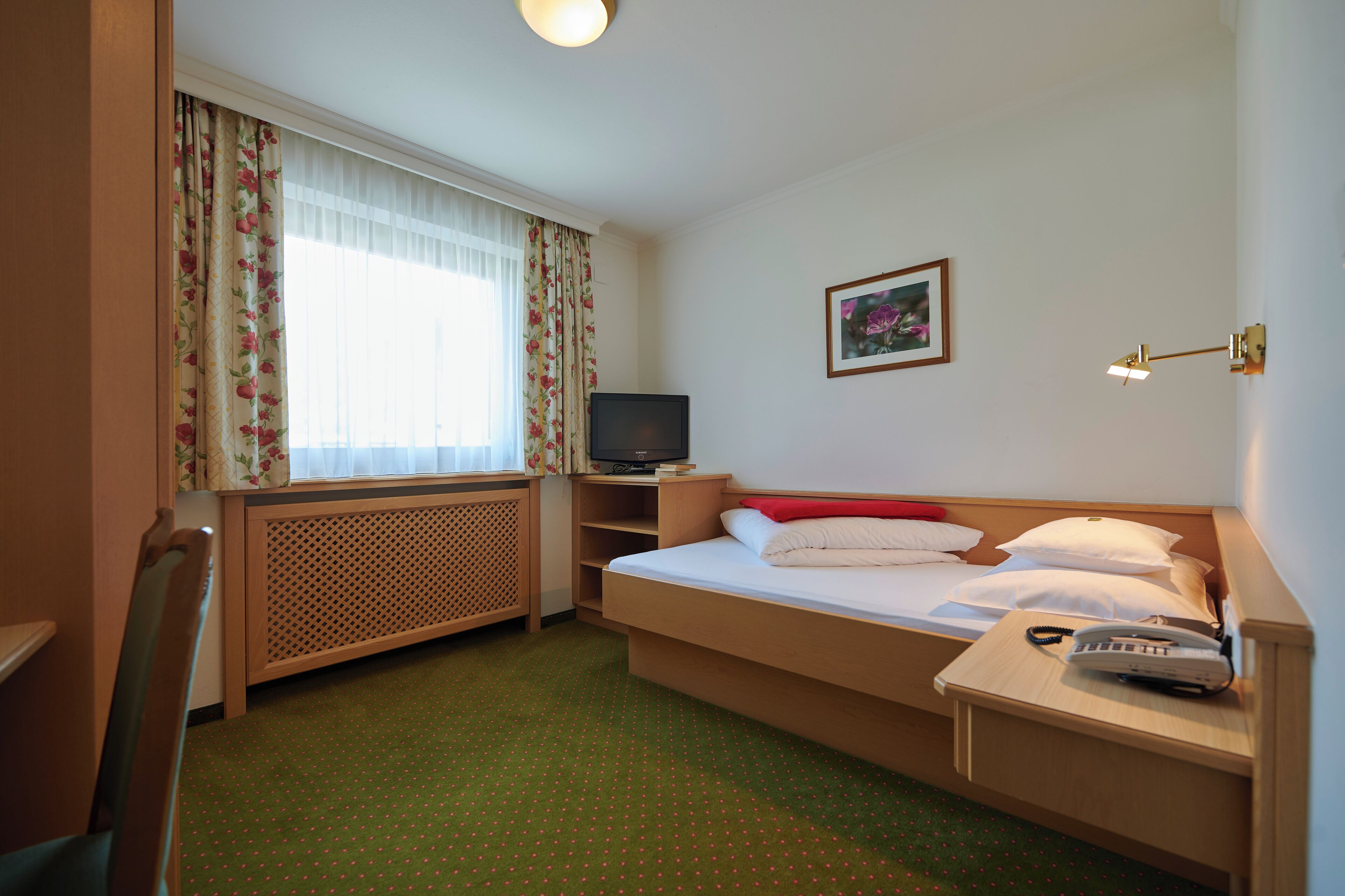 Habitación de hotel con cama, escritorio, silla, televisor, teléfono y ventana con cortinas.