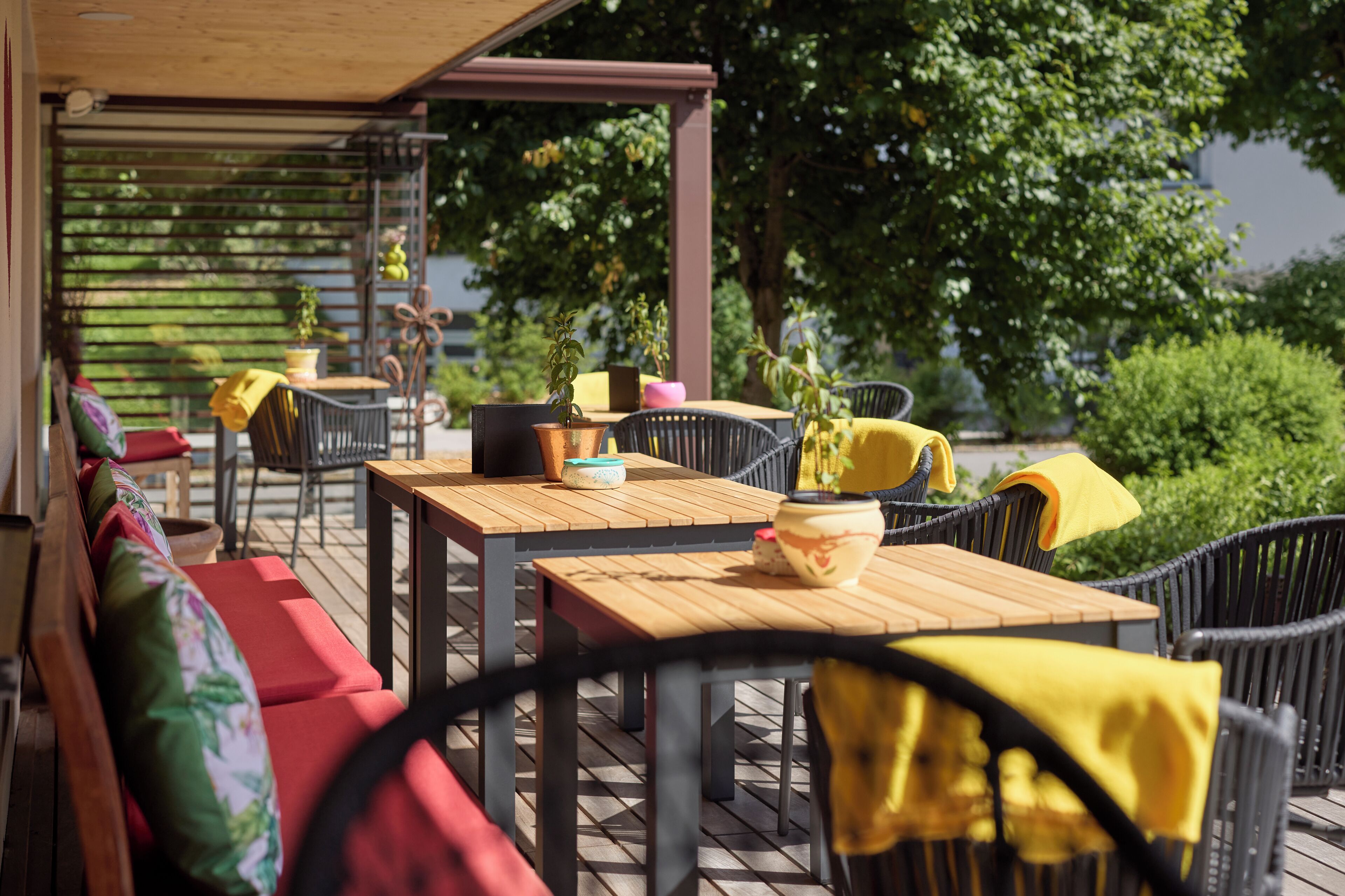 An outdoor patio with wooden tables and chairs, some with yellow cloths. There are potted plants and a wooden pergola structure.