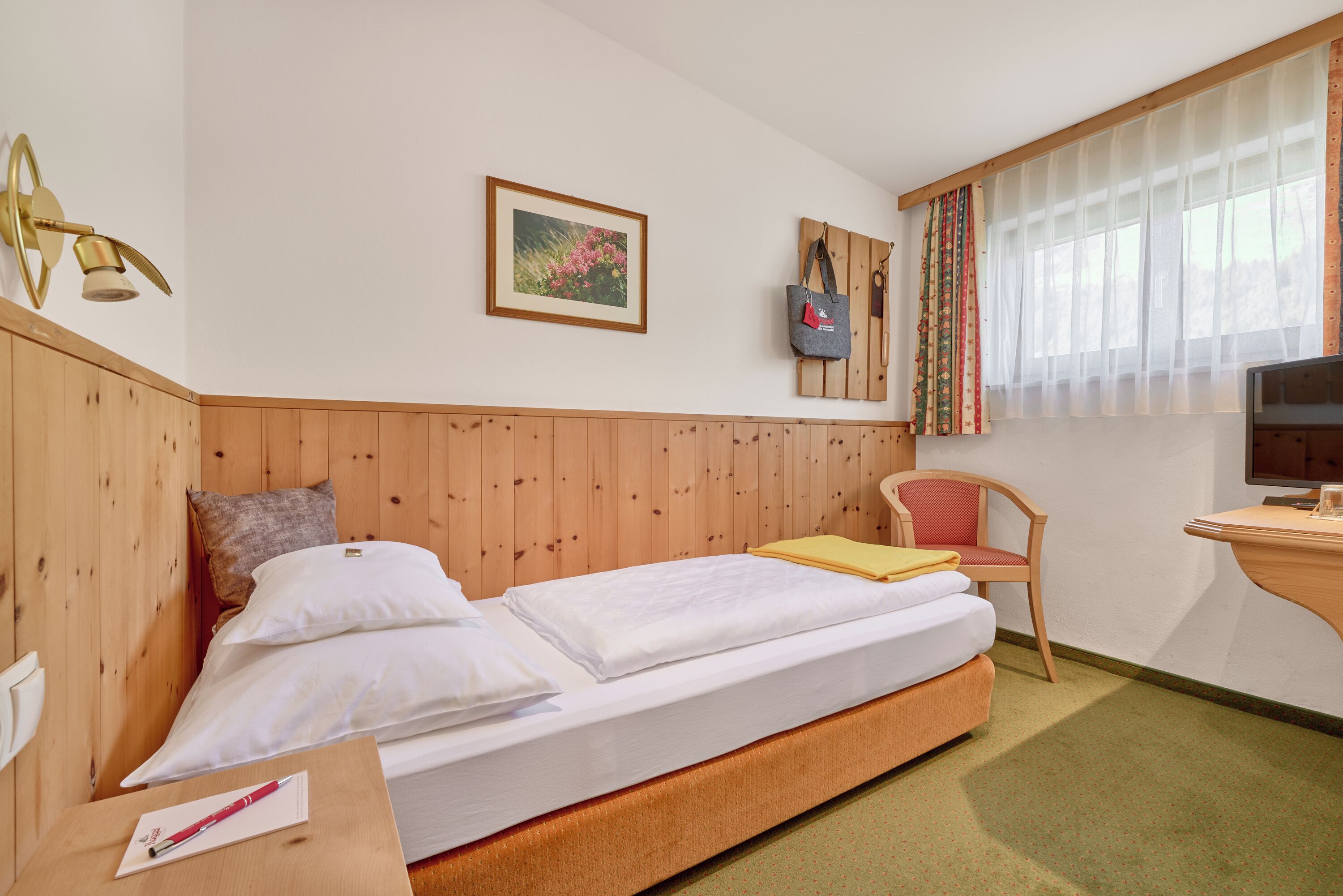 Ett hotellrum med en sänggavel i trä, en säng med vita sängkläder, ett skrivbord med en stol, en tv och ett fönster med tunna, genomskinliga gardiner.