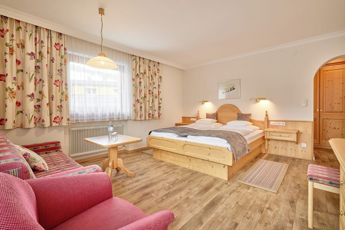 A cozy bedroom with a wooden bed, a small bedside table, a chair, and a window with floral curtains.