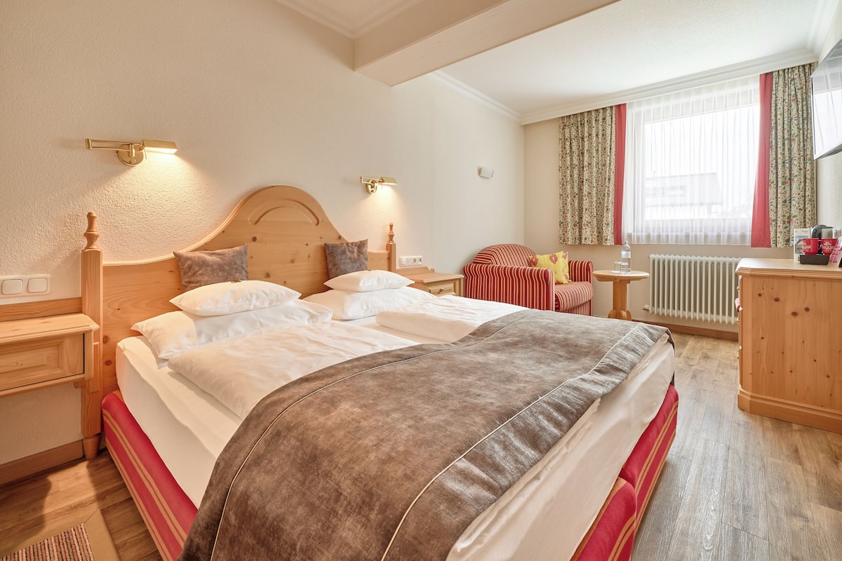 A hotel room with a large bed, wooden headboard, bedside tables, a nightstand, a dresser, a window with curtains, and a radiator.
