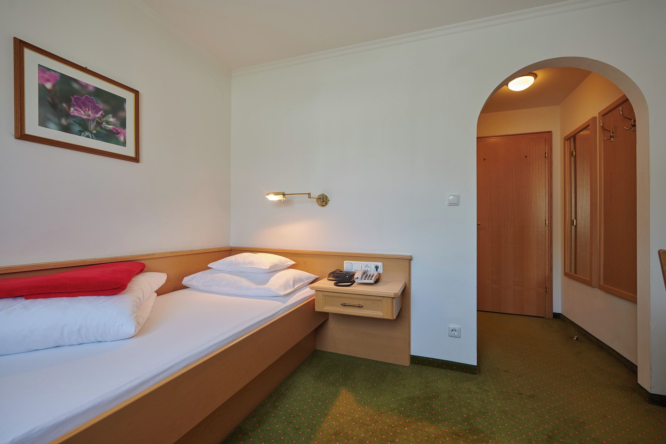 Habitación de hotel con cama, mesita de noche con lámpara, un cuadro enmarcado en la pared y un pasillo que conduce a una puerta.