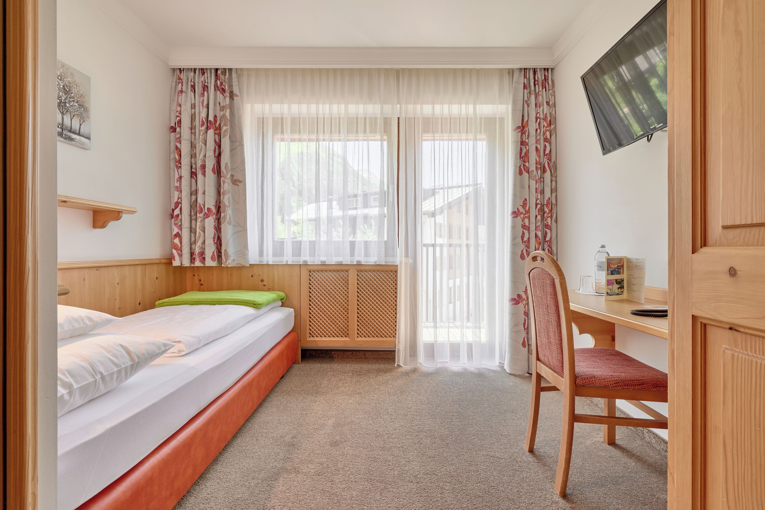 Un dormitorio con cama, escritorio, silla, televisor y ventana con cortinas.