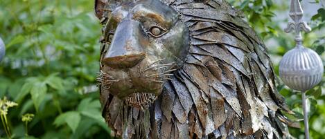A metal sculpture of a lion's head with a textured mane, set against a backdrop of greenery.