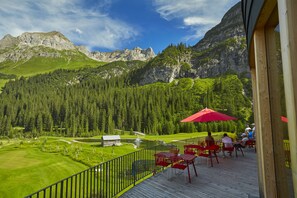 A deck with red chairs and tables overlooking a golf course and mountains. - Hotel Appartement Roggal (Lech am Arlberg)