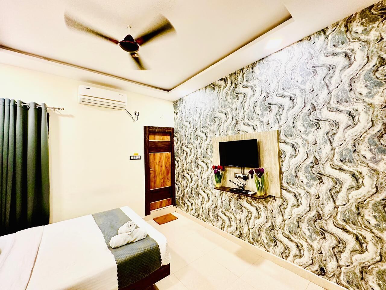Habitación de hotel con cama, ventilador de techo, unidad de aire acondicionado, televisor montado en una pared con diseño ondulado y una pequeña estantería con flores.