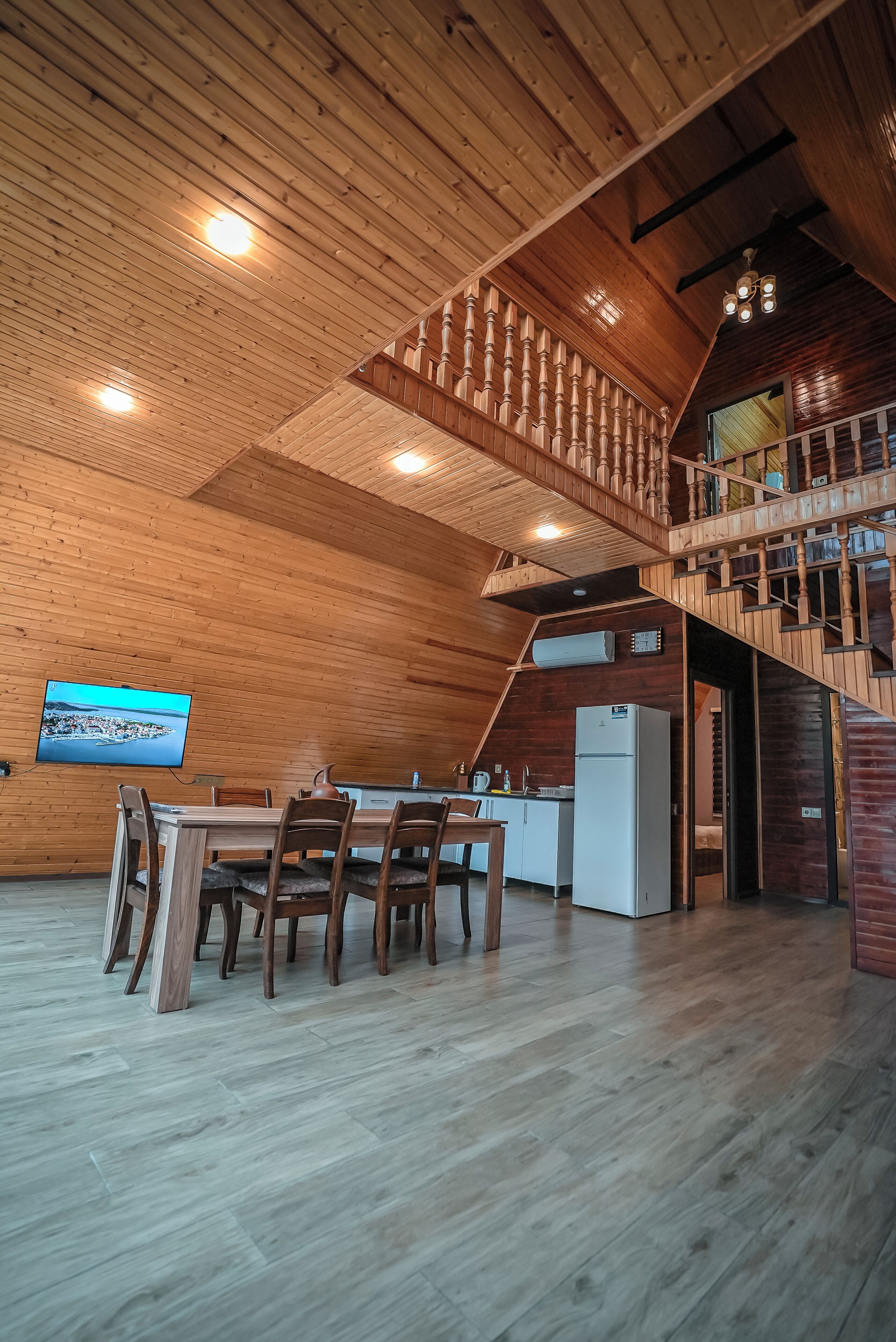 A spacious room with a wooden ceiling, a dining area with a table and chairs, a kitchen area with a refrigerator, and a staircase leading to an upper level.