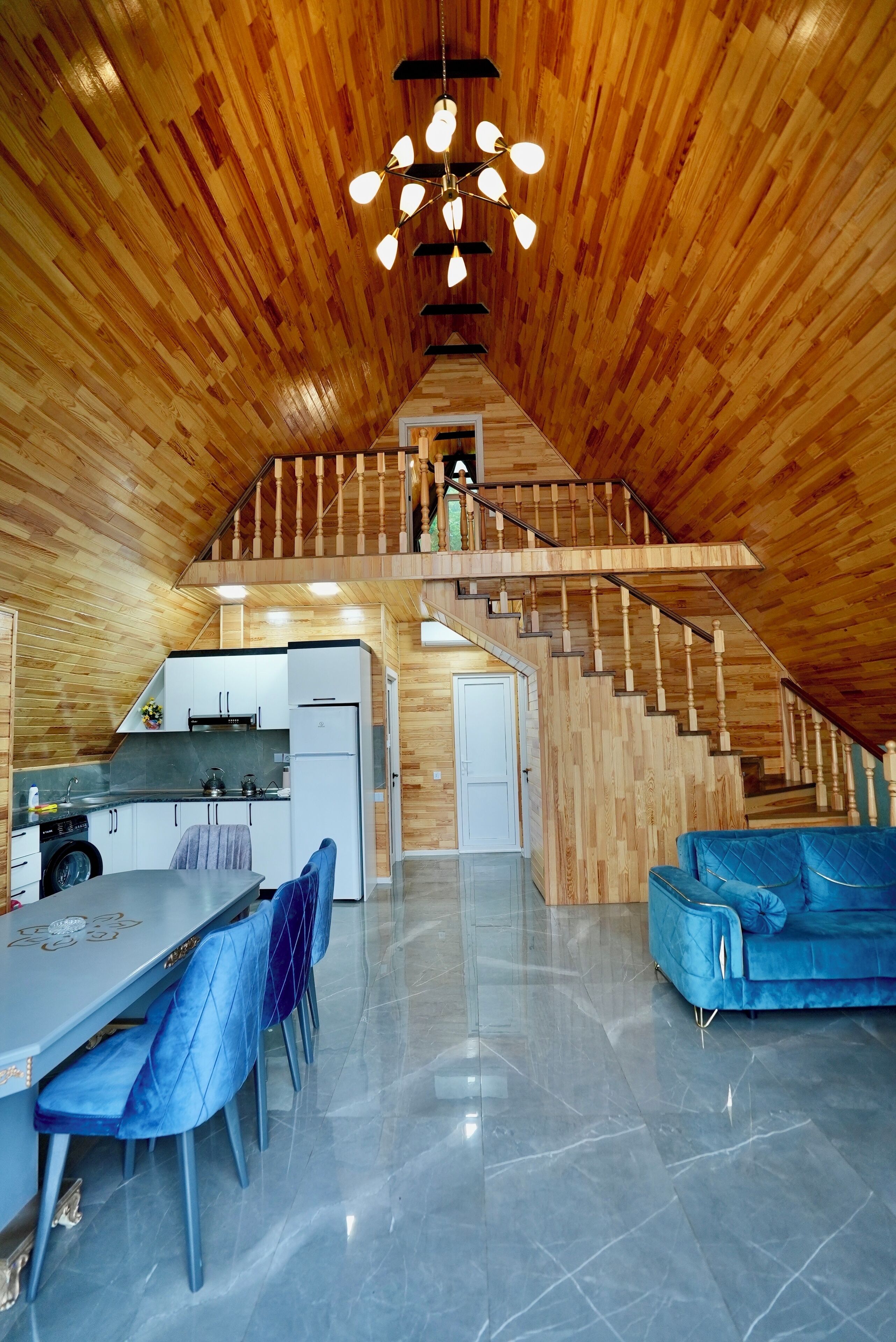 A spacious loft with a wooden interior, a dining area with blue chairs, a kitchen, and a staircase leading to an upper level.