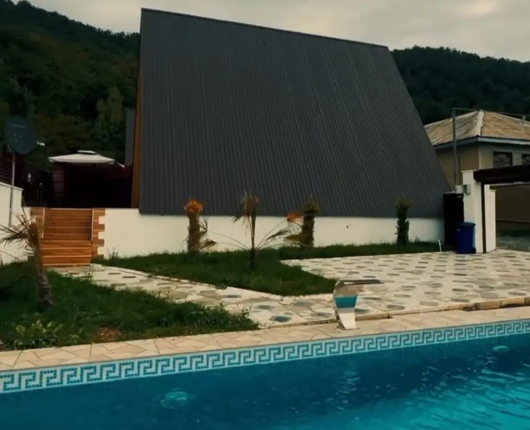 A modern building with a large sloped roof, a swimming pool with a decorative edge, and a paved patio area.