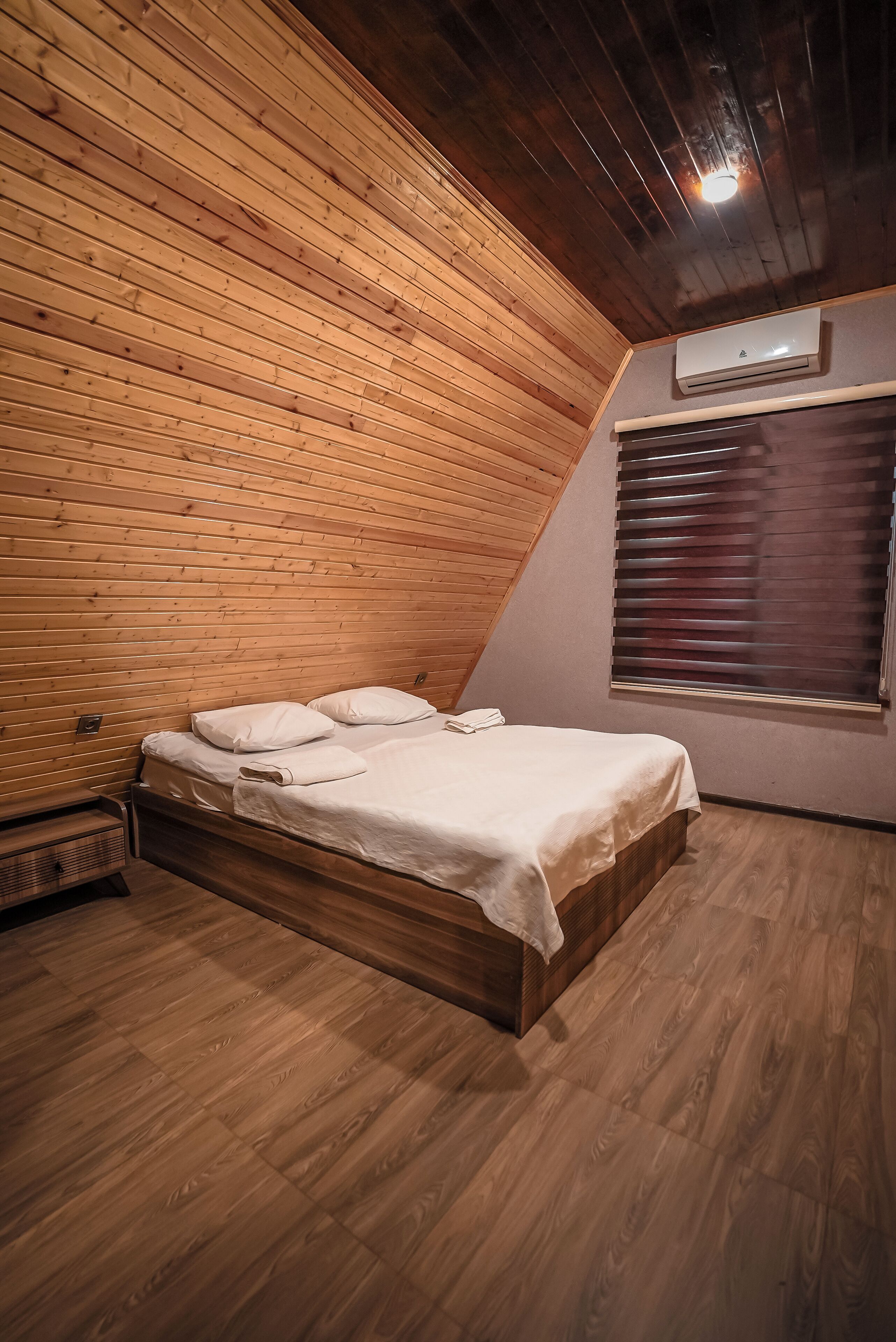 A room with a wooden sloped ceiling, a bed with white bedding, a window with blinds, and an air conditioning unit.