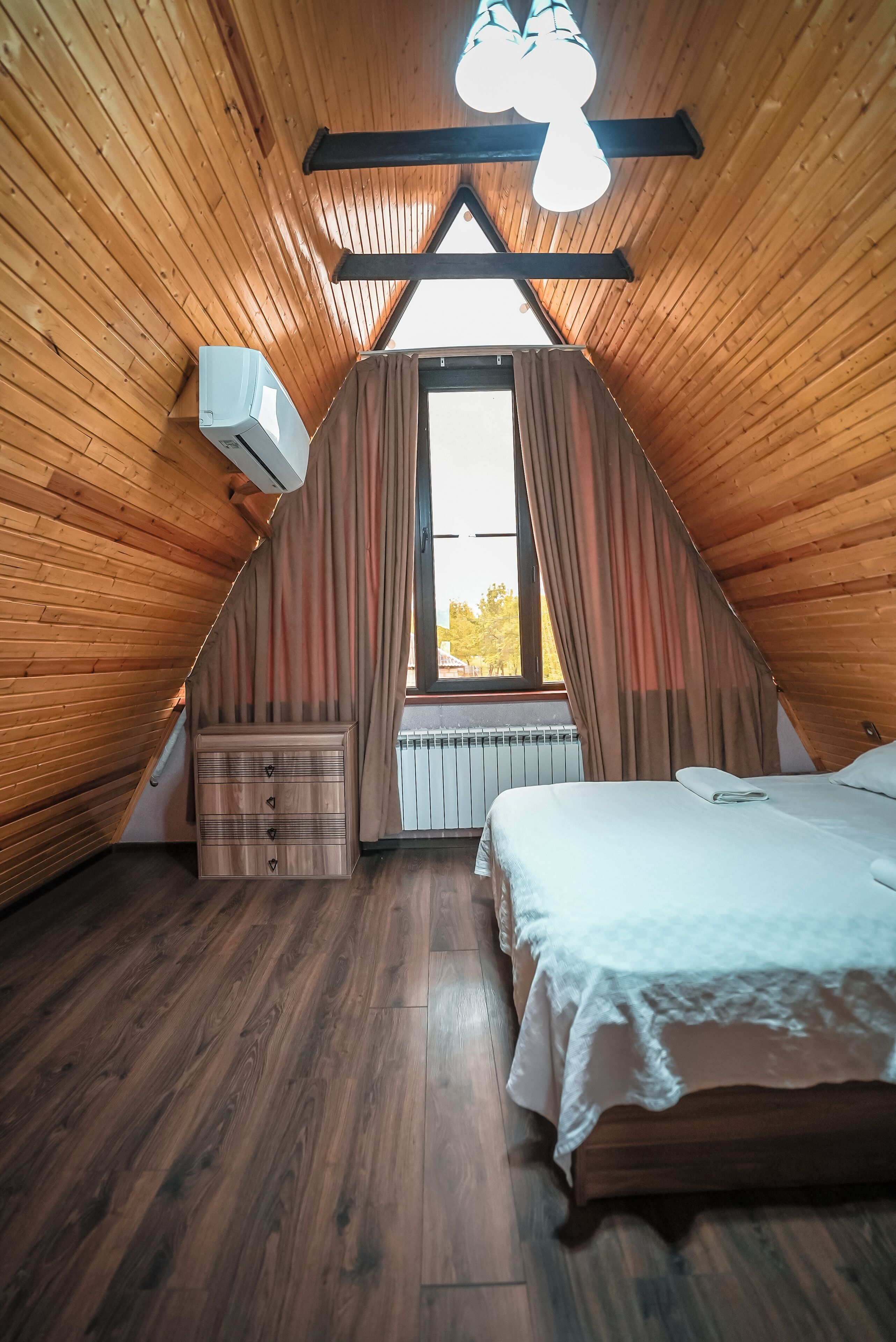 A cozy attic bedroom with wooden walls, a large window with curtains, a bed, a dresser, and an air conditioning unit.