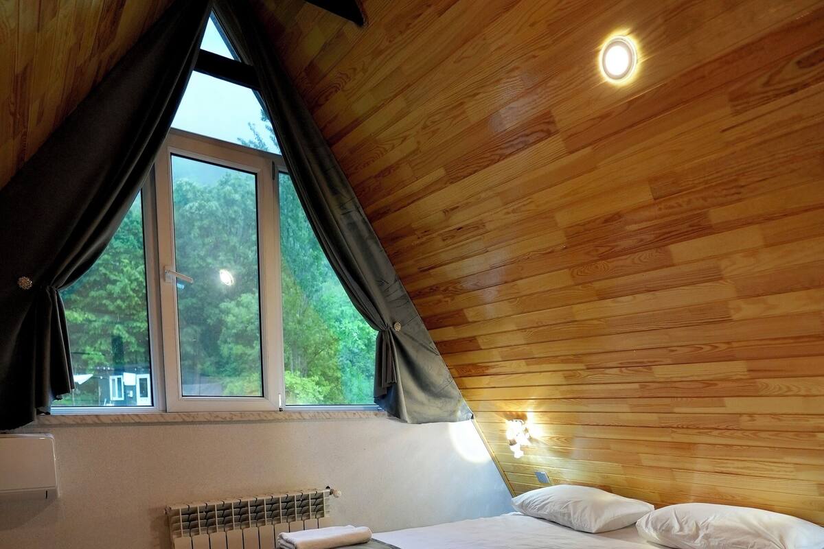A bedroom with a wooden ceiling, a bed with white linens, a window with curtains, and a wall-mounted light.