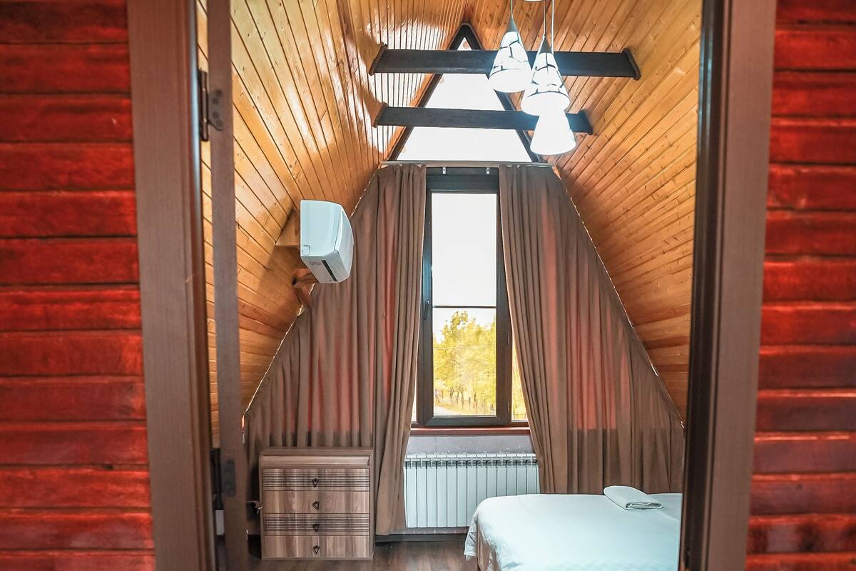 A bedroom with a sloped ceiling, wooden flooring, a bed, a nightstand, a dresser, and a window with curtains.