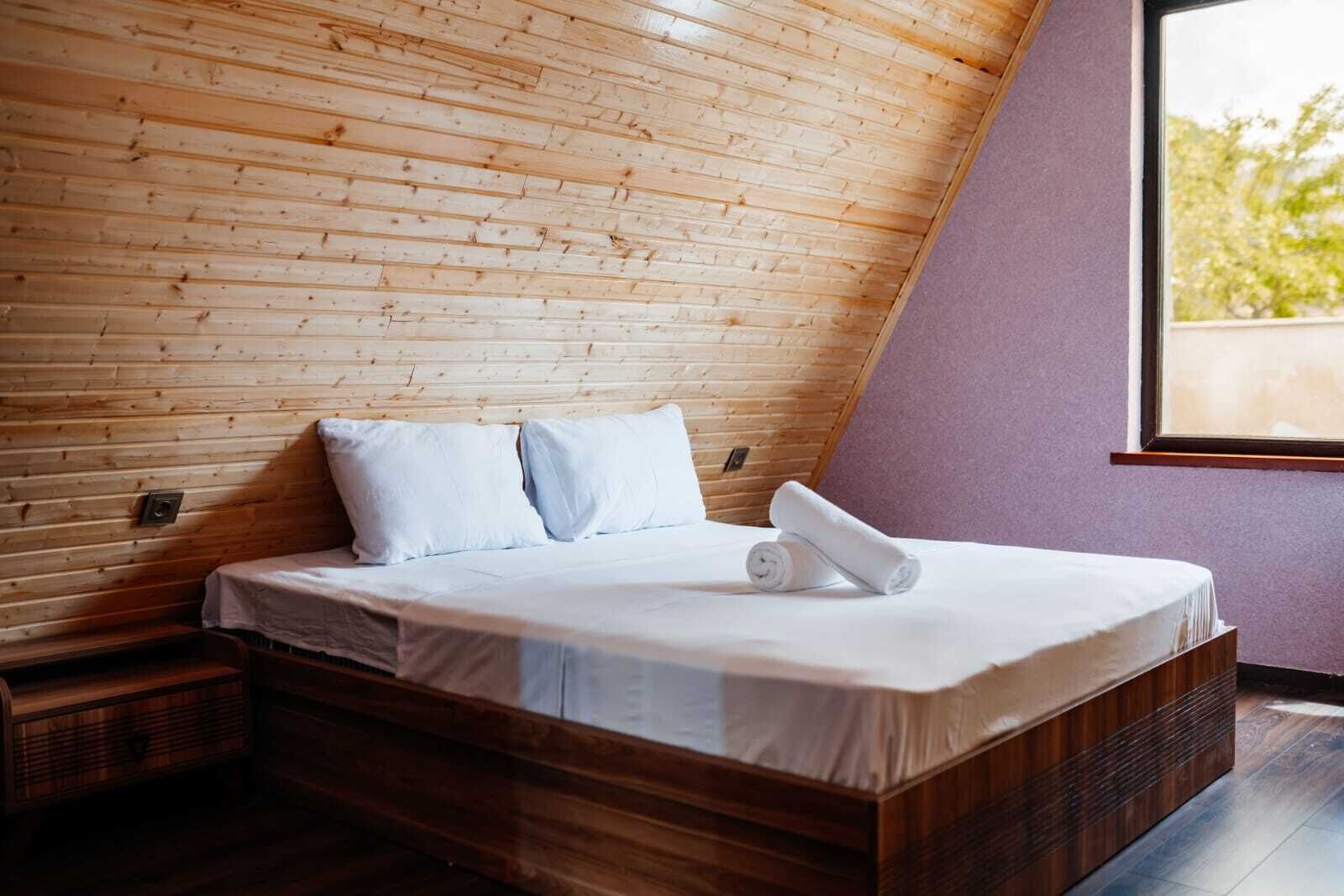 A bedroom with a wooden sloped ceiling, a bed with white linens, and a window with a view of trees.