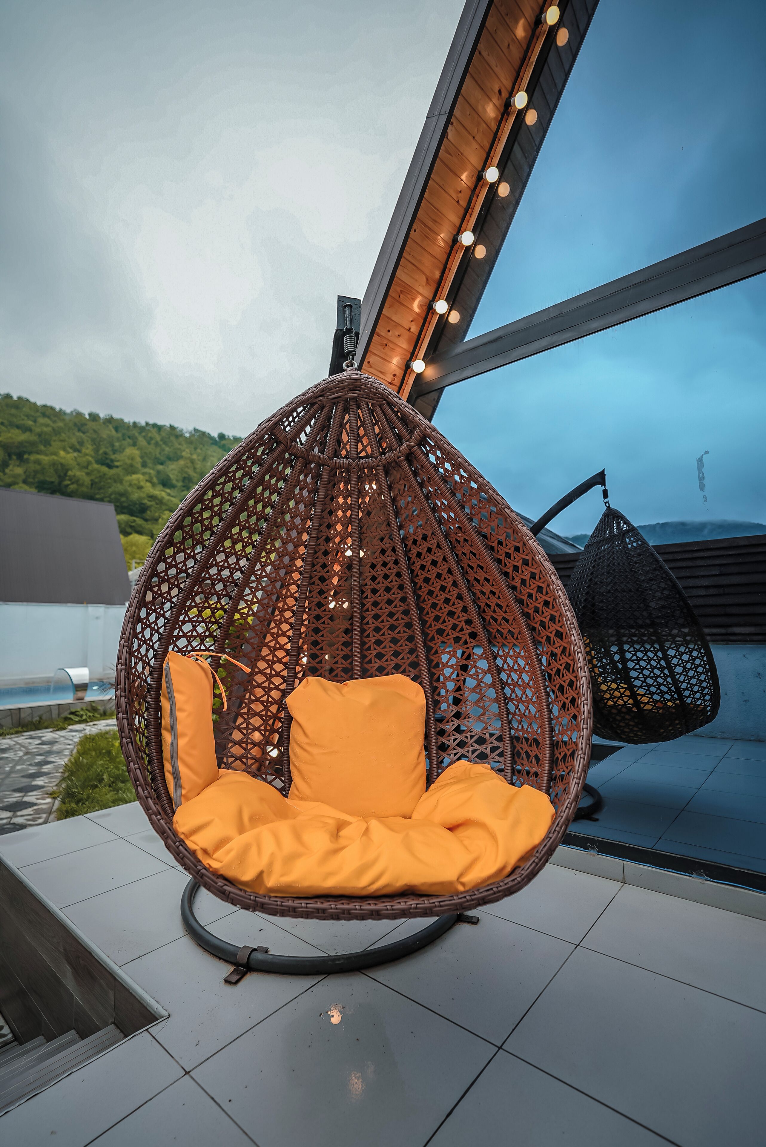 A wicker swing chair with orange cushions on a tiled patio overlooking a body of water and greenery.