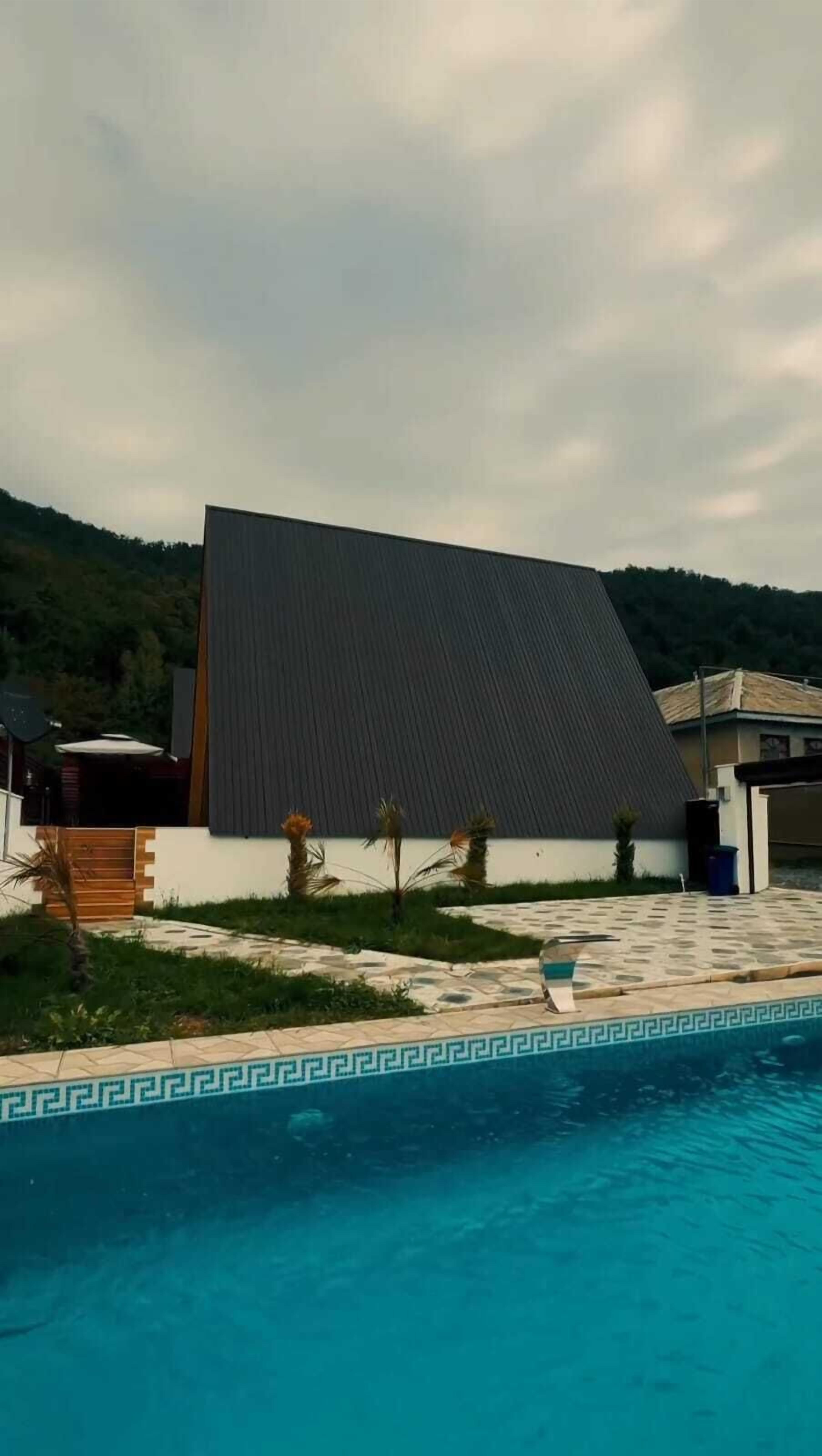 Ein modernes Haus mit einem großen, geneigten Dach, einem Swimmingpool mit dekorativem Rand und einem Gartenbereich mit kleinen Palmen.