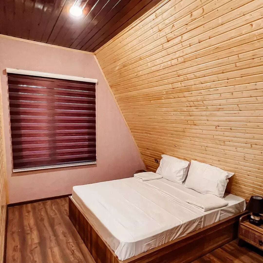 Ein Schlafzimmer mit schräg steilender Decke, Holzwänden, einem Bett mit weißer Bettwäsche und einem Fenster mit heruntergelassenen Jalousien.
