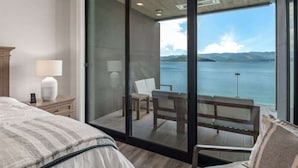 A bedroom with a large bed, a bedside table with a lamp, and a view of a balcony with a table and chairs.