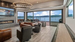 A spacious living area with a fireplace, comfortable seating, and large windows offering a view of a lake and mountains.