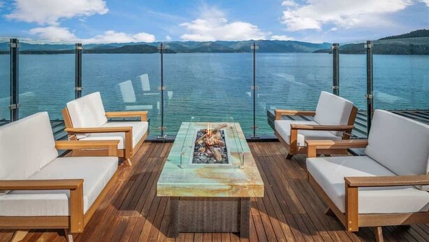 A deck with wooden chairs and a central fire feature, overlooking a calm lake with distant mountains.