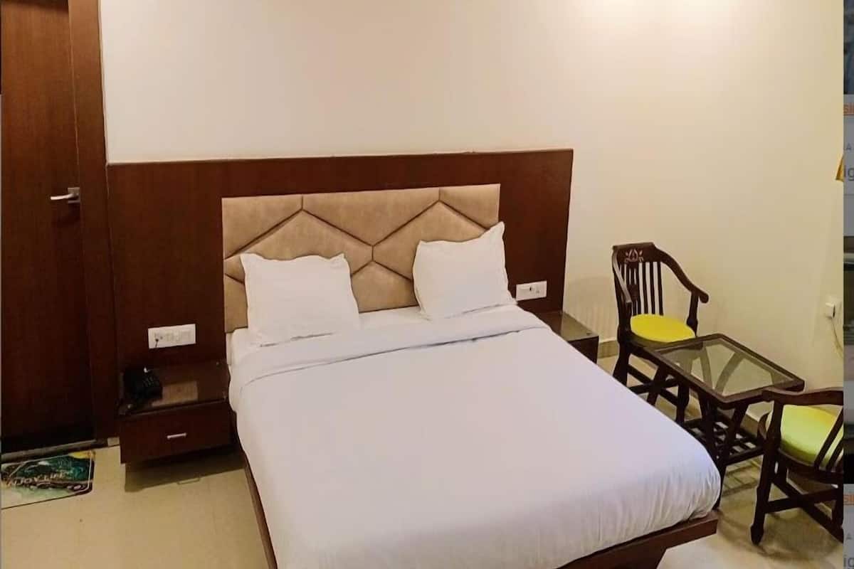 Uma cama de solteiro com cabeceira de madeira e duas almofadas, uma pequena mesa de madeira com uma cadeira, e uma porta à esquerda.
