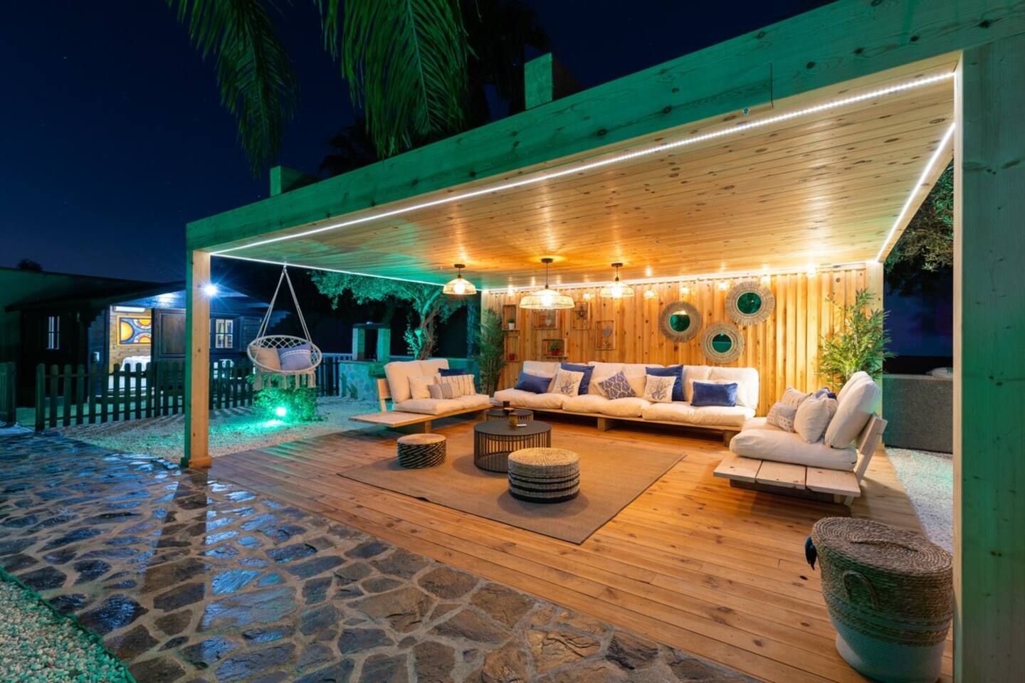 An outdoor patio with a wooden deck, a sofa set, hanging chairs, and a stone-tiled floor.