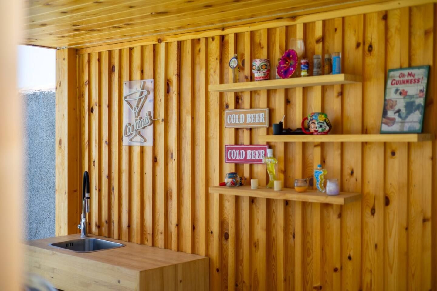 Un intérieur en bois avec un évier, des étagères remplies de divers objets et des panneaux indiquant « BIÈRE FROIDE ».