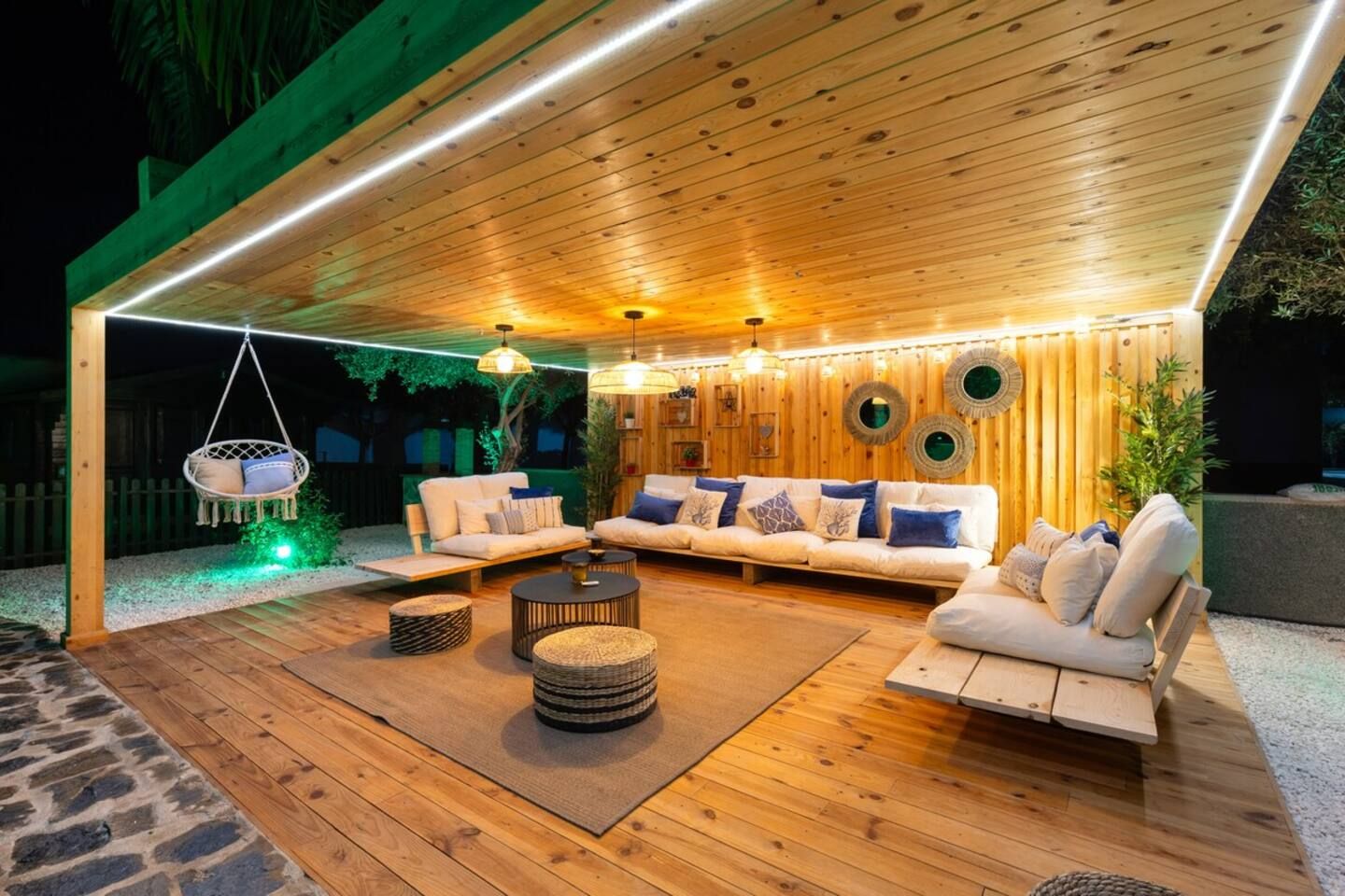 An outdoor patio with wooden flooring, a sofa set, hanging chairs, and ambient lighting.