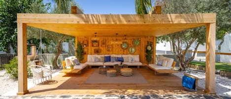 An outdoor wooden structure with a sofa and chairs, surrounded by palm trees and a stone floor.