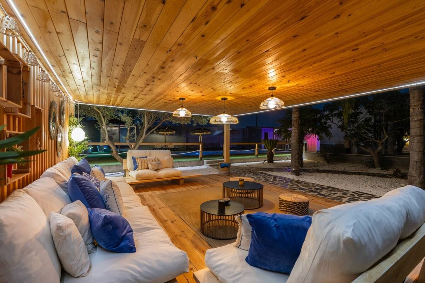 An outdoor seating area with wooden flooring, cushioned sofas, and round tables.