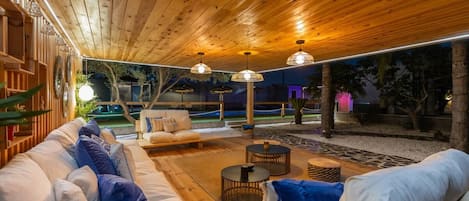 An outdoor seating area with wooden flooring, cushioned sofas, and round tables.